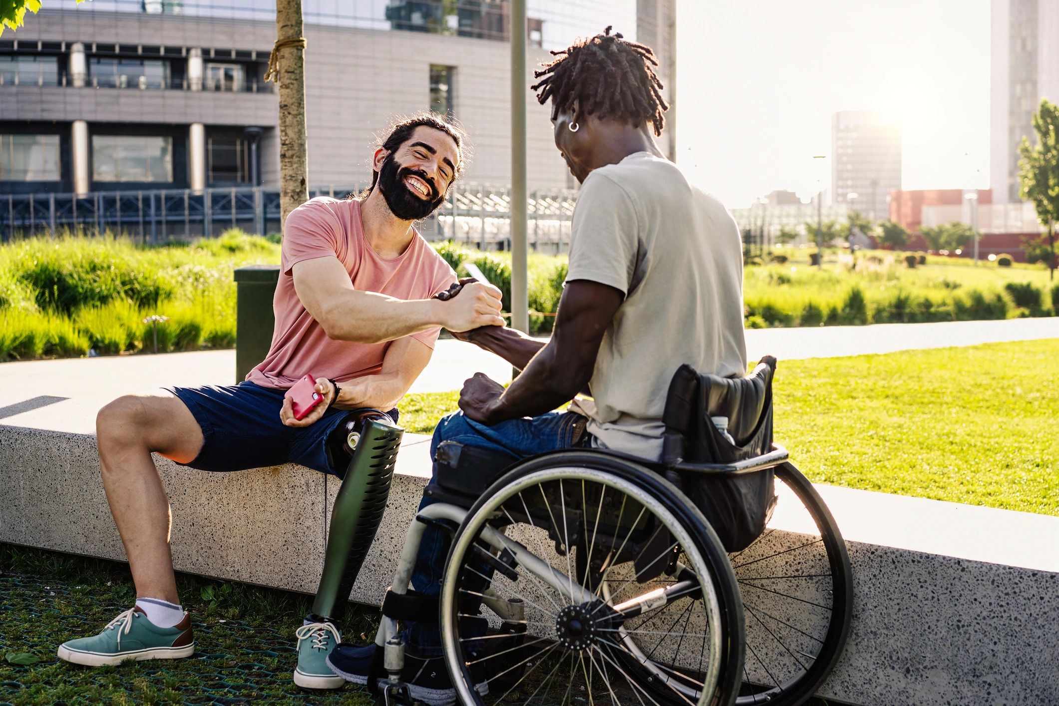Two men, one with a prosthetic leg and one in a wheelchair, share a joyful handshake outdoors.
